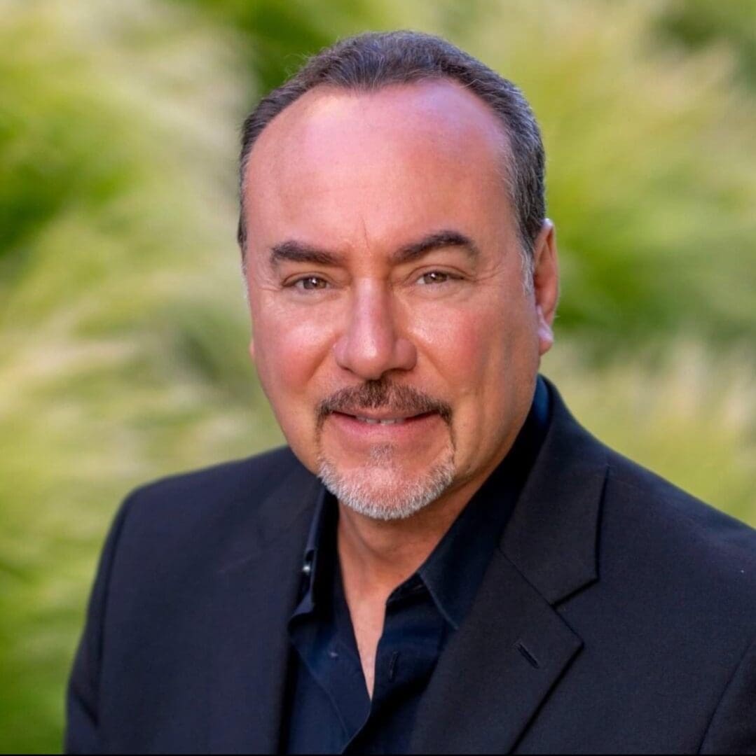Smiling man in a black suit outdoors with greenery in the background.